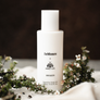 A bottle of Aro Ha X Arithmos Superfine Body Oil with a white label, placed on a surface with greenery in the background.